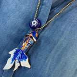 Ballchain necklace with a blue and gold enamel fish pendant and evil eye amulet on a denim shirt background.