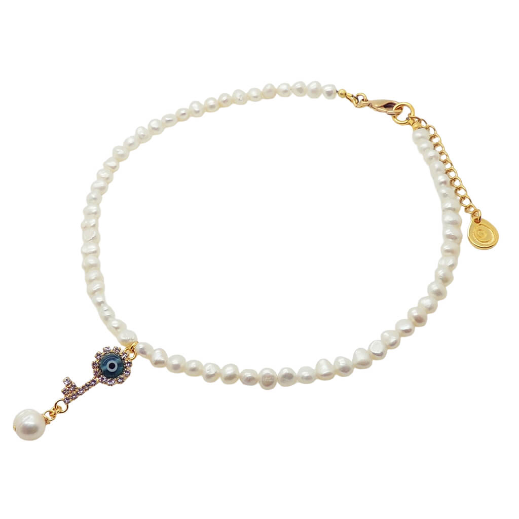 A Dana Levy white freshwater pearl choker with a gold plated clasp and extender chain, featuring a dangling diamante rhinestone evil eye key charm with a blue glass evil eye centre, on a white background.