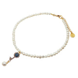 A Dana Levy white freshwater pearl choker with a gold plated clasp and extender chain, featuring a dangling diamante rhinestone evil eye key charm with a blue glass evil eye centre, on a white background.