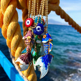 Colourful enamel fish and evil eye amulet chain necklaces hanging from a rope with a blurred ocean background
