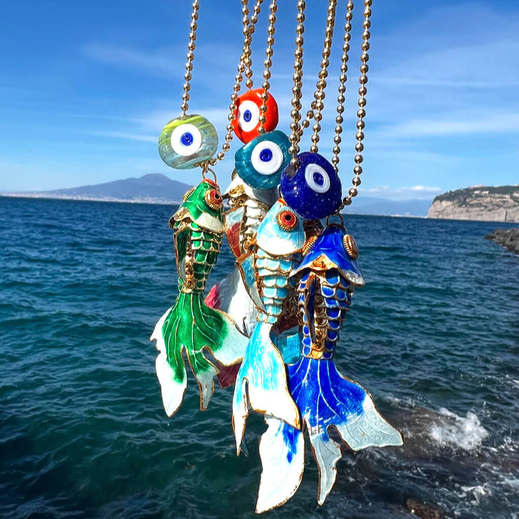 Colourful enamel fish and evil eye charm pendants on ballchain necklaces against a backdrop of ocean and sky.