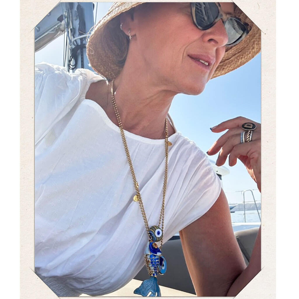 Model wearing a straw hat, sunglasses, and a white shirt with a blue enamel fish and evil eye pendant necklace on a boat.