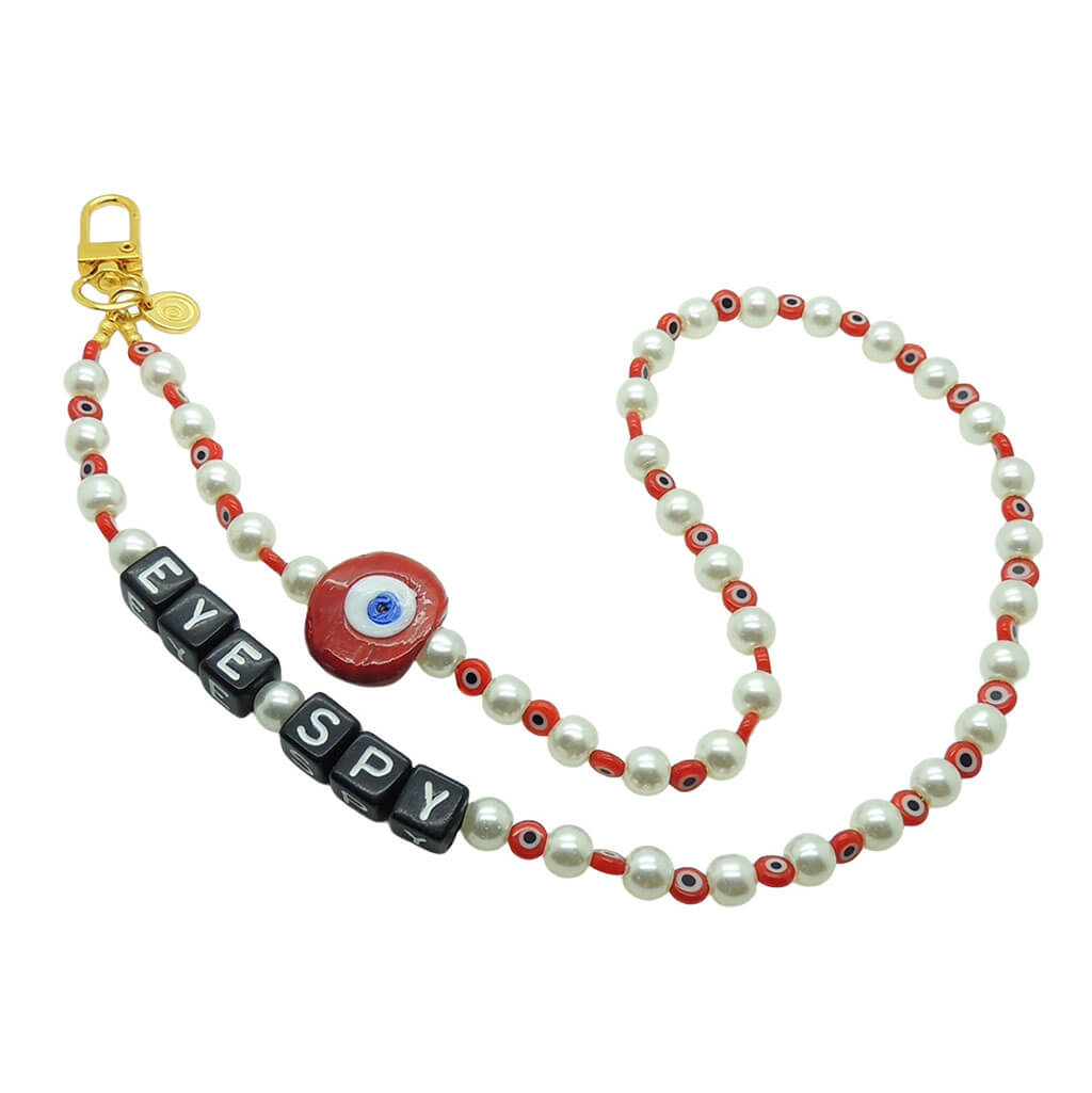 Dana Levy personalised faux pearl phone charm strap with red evil eye charm and beads and black alphabet cube 'EYE SPY' beads, with a gold plated clasp on a white background.