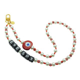 Dana Levy personalised faux pearl phone charm strap with red evil eye charm and beads and black alphabet cube 'EYE SPY' beads, with a gold plated clasp on a white background.