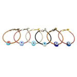 Dana Levy six braided leather cord bracelets with chunky glass evil eye amulets, displayed in a row against a plain white background. The cords are in tan brown, pink, white, and black tones, and the evil eye amulets are in various shades of blues and white.