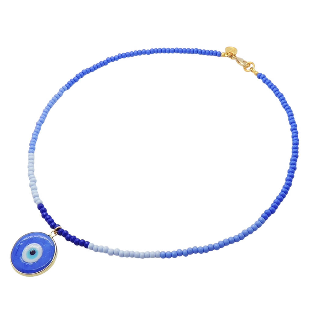 A glass beaded necklace by Dana Levy featuring blue and light blue glass rocaille beads with a flat round blue evil eye pendant, finished with a gold clasp, shown on a plain white background.