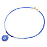 A glass beaded necklace by Dana Levy featuring blue and light blue glass rocaille beads with a flat round blue evil eye pendant, finished with a gold clasp, shown on a plain white background.