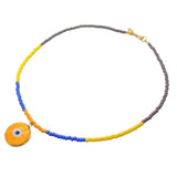 A Dana Levy glass bead necklace with a round orange evil eye disc pendant, featuring grey, yellow, blue and orange beads, and a gold clasp on a white background.