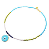 A Dana Levy glass beaded necklace with a light blue and turquoise evil eye pendant, featuring segments of turquoise blue, olive green, baby pink, and dark navy blue beads, finished with a gold clasp and charm.