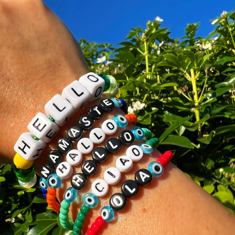 A close-up of a person's wrist wearing multiple colorful beaded bracelets outdoors, with green foliage and blue sky in the background. The bracelets include white letter beads spelling out 'HELLO' and 'NAMASTE', black letter beads spelling 'HALLOO', and several bright multicolor bead strands.