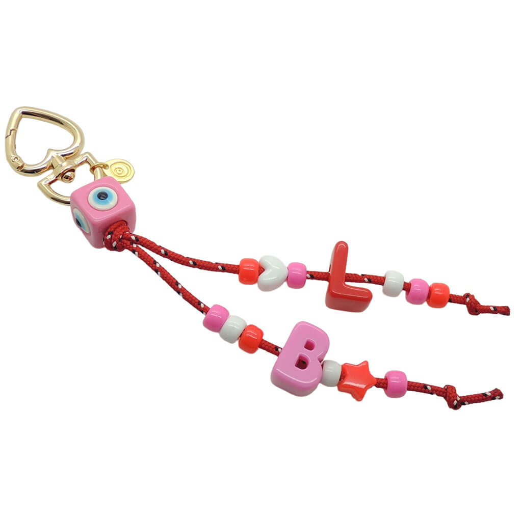 A Dana Levy maxi alphabet initial bag charm with red nautical cords, a pink evil eye cube charm, L and B maxi alphabet letters and mini charms on a white background. 