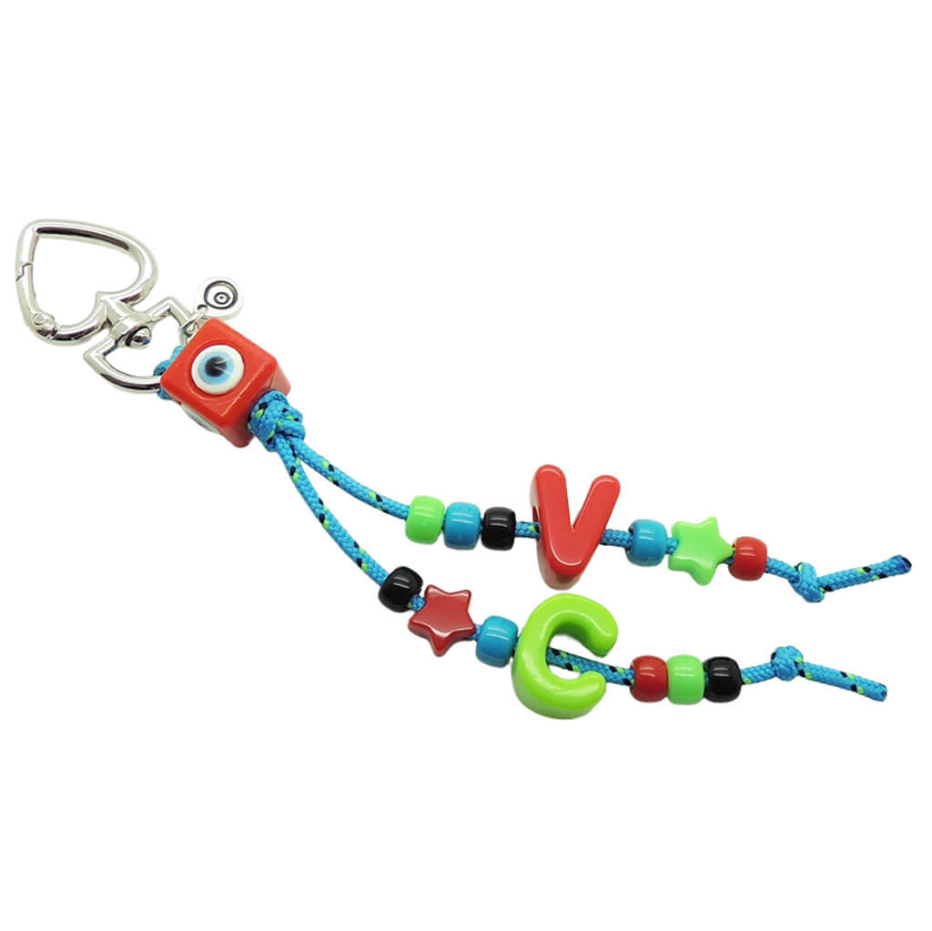 A Dana Levy maxi alphabet initial bag charm with turquoise cords, a red evil eye cube charm, V and C maxi alphabet letters and mini charms on a white background. 