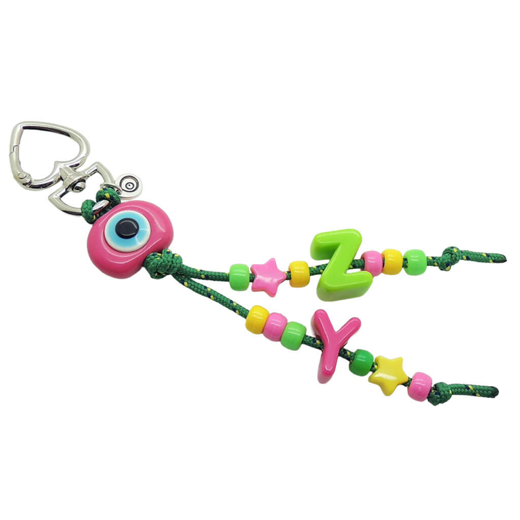 A Dana Levy maxi alphabet initial bag charm with green cords, a magenta evil eye dome charm, Z and Y maxi alphabet letters and mini charms on a white background. 