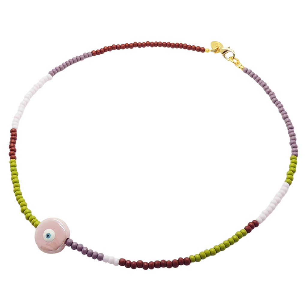 A glass bead necklace by Dana Levy with brown, purple, lilac and olive glass beads, and a large flat pearlised pink ceramic evil eye disc pendant in the centre, on a white background. 