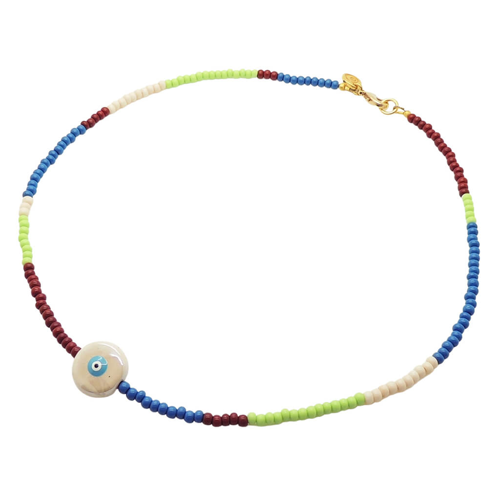 A Dana Levy glass bead necklace with alternating sections of blue, pistachio green, cream, and brown beads, featuring a round ceramic creme coloured evil eye charm and a gold clasp, shown on a plain white background.