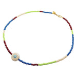 A Dana Levy glass bead necklace with alternating sections of blue, pistachio green, cream, and brown beads, featuring a round ceramic creme coloured evil eye charm and a gold clasp, shown on a plain white background.