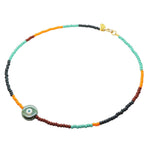 A glass bead necklace by Dana Levy with segments of aqua, forest green, orange, and brown beads, featuring a round ceramic pearlised forest green and white evil eye charm and a gold clasp, shown on a plain white background.