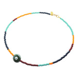 A Dana Levy beaded necklace with a multicolour sequence of aqua, dark green, orange, and burgundy glass beads, featuring a central ceramic forest green and white evil eye bead and a gold clasp, shown on a plain white background.