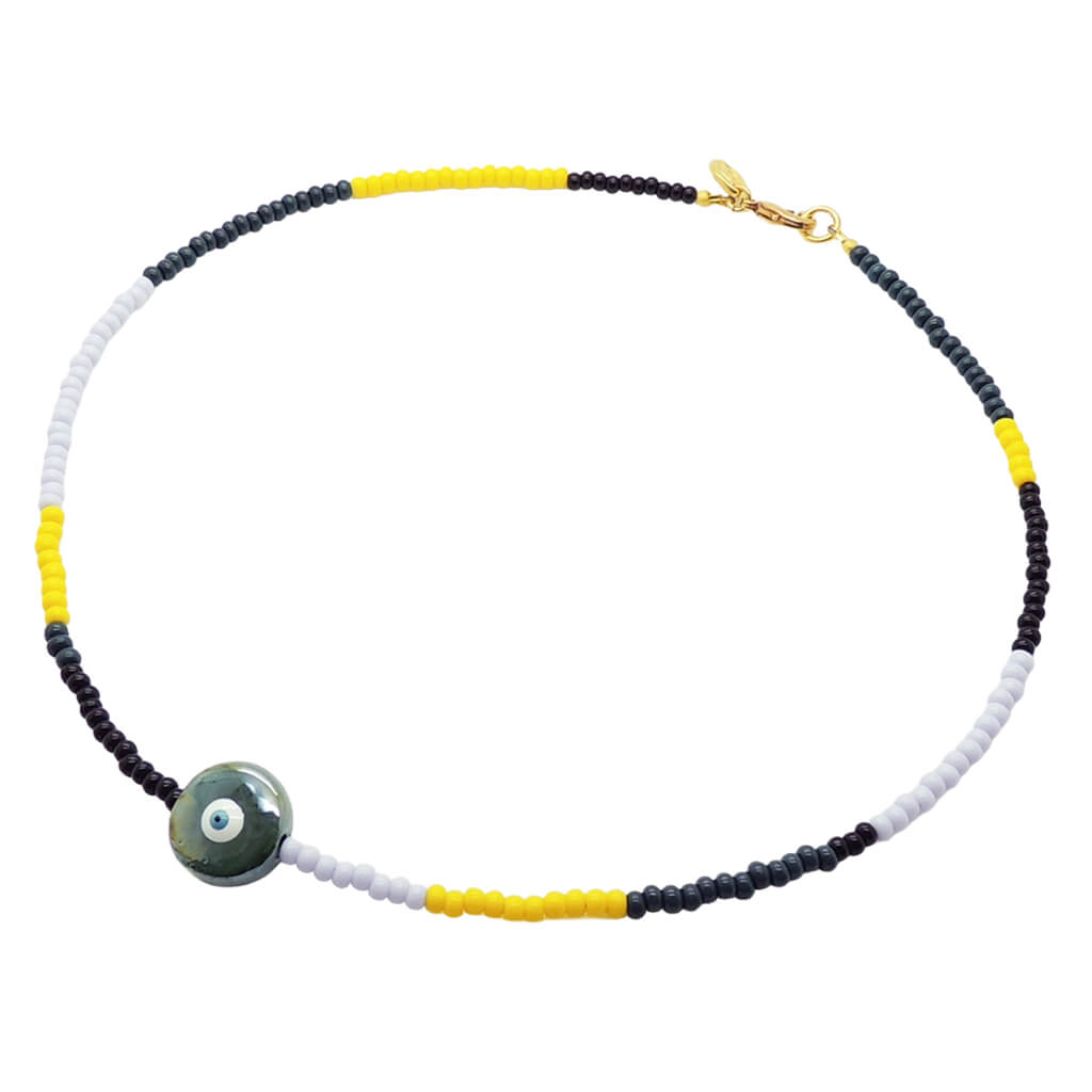 A Dana Levy glass bead necklace with black, yellow, forest green and lilac glass beads, and a pearlised forest green ceramic evil eye disc pendant in the centre, on a white background. 