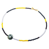 A Dana Levy glass bead necklace with black, yellow, forest green and lilac glass beads, and a pearlised forest green ceramic evil eye disc pendant in the centre, on a white background. 
