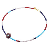 A Dana Levy beaded necklace with repeating segments of navy blue, light pink, turquoise, and red glass beads, featuring a large ceramic pearlised maroon evil eye charm and a gold clasp, shown on a plain white background.