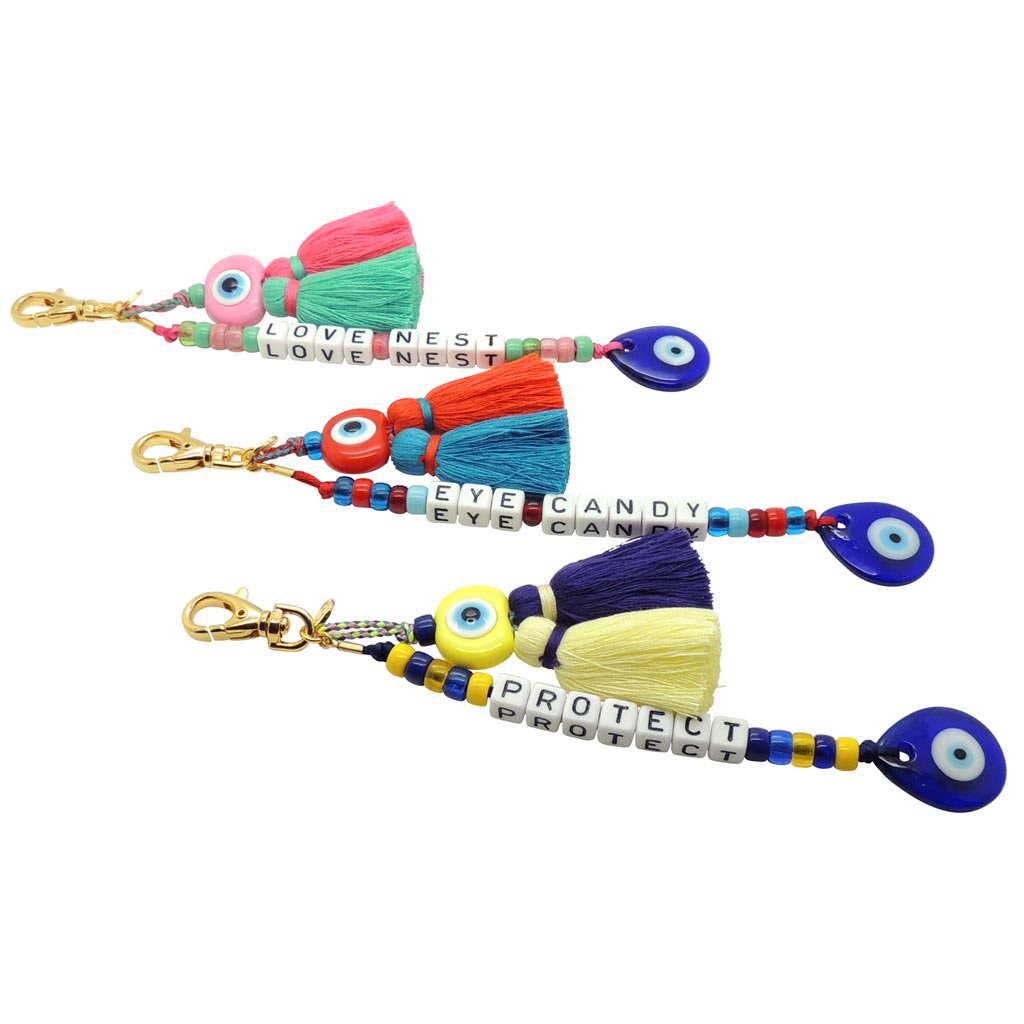Dana Levy Personalised Alphabet Cube Evil Eye Charm Tassel Keyring - A Trio of Gold Plated Keyrings with Love Nest, Eye Candy and Protect Written with Alphabet Letter Cubes and Decorated with Evil Eye Charms and Colourful Tassels.