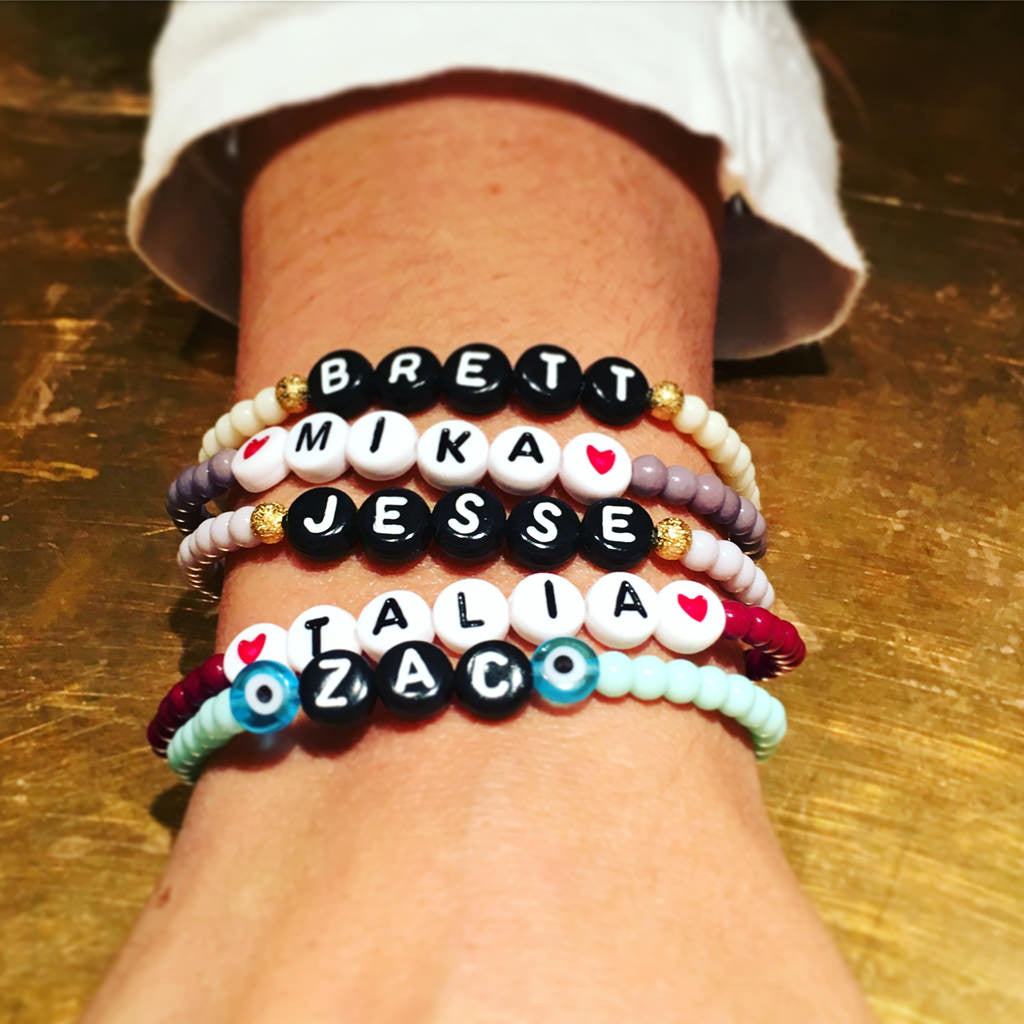 Dana Levy five personalised name bead bracelets with a variety of names and bead colours on wrist on a gold background.